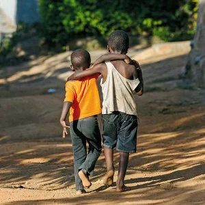 Zwei schwarze, barfüßig gehende, Kinder, Schulter an Schulter auf einem Sandweg